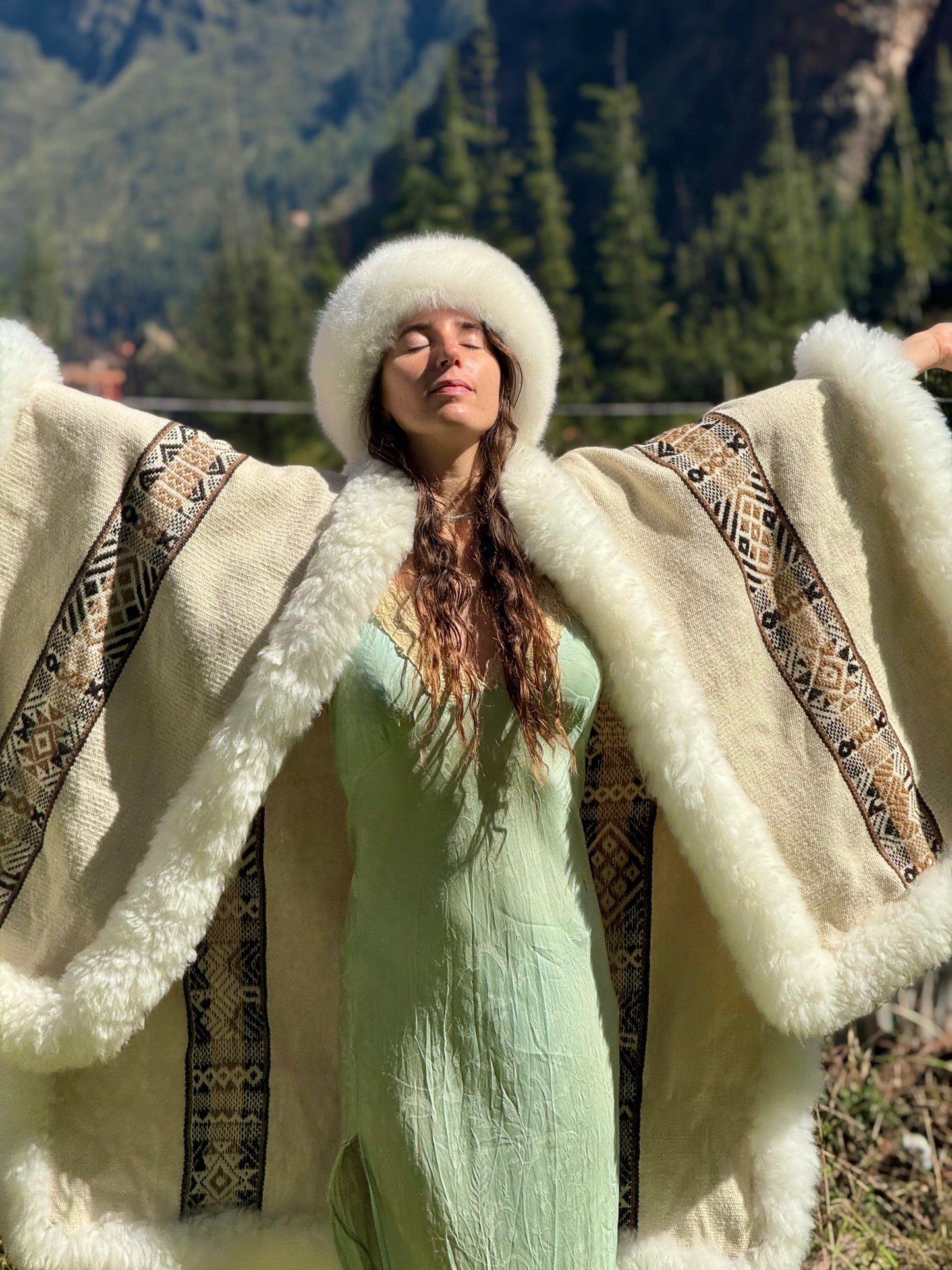 Woman in a green dress with a white fur coat and hat, standing in a mountainous area.